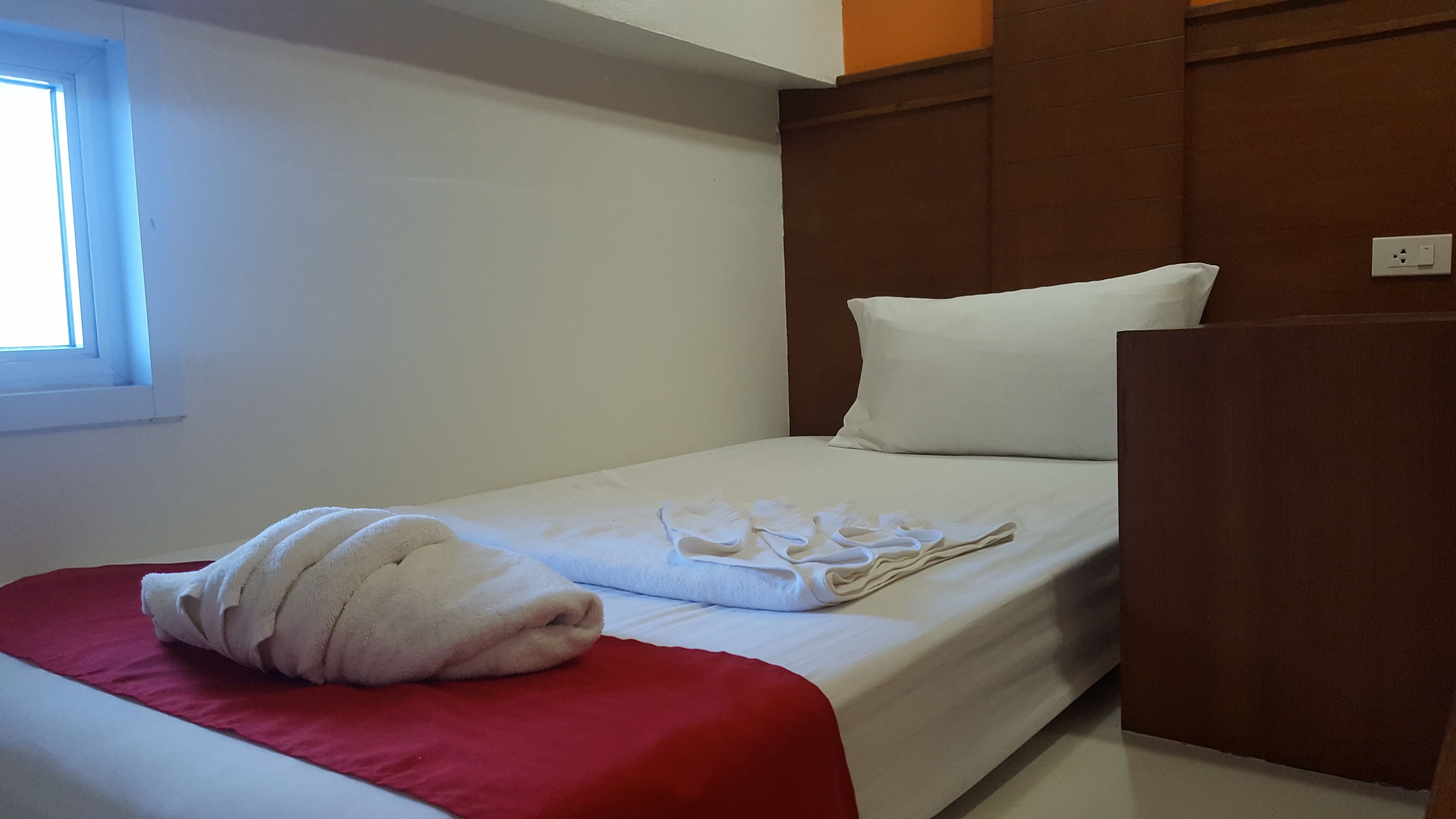 A single bed with a red blanket, white pillows, and a rolled white towel.