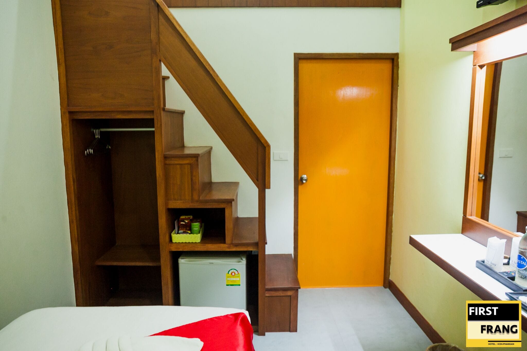 A hotel room with a wooden staircase, a small refrigerator, a bed with a red and white bedspread, and a desk with a mirror and a tray.