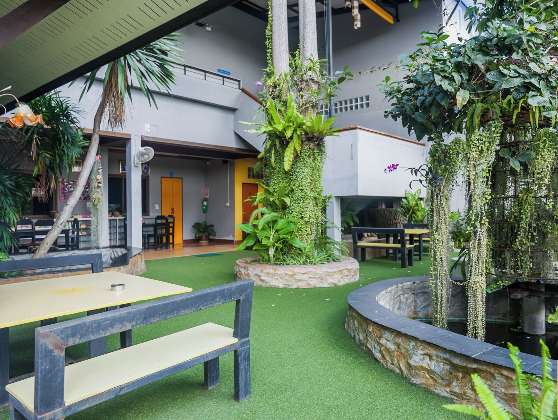 Jardí interior amb gespa artificial, bancs i una font rodada per una vegetació abundant.