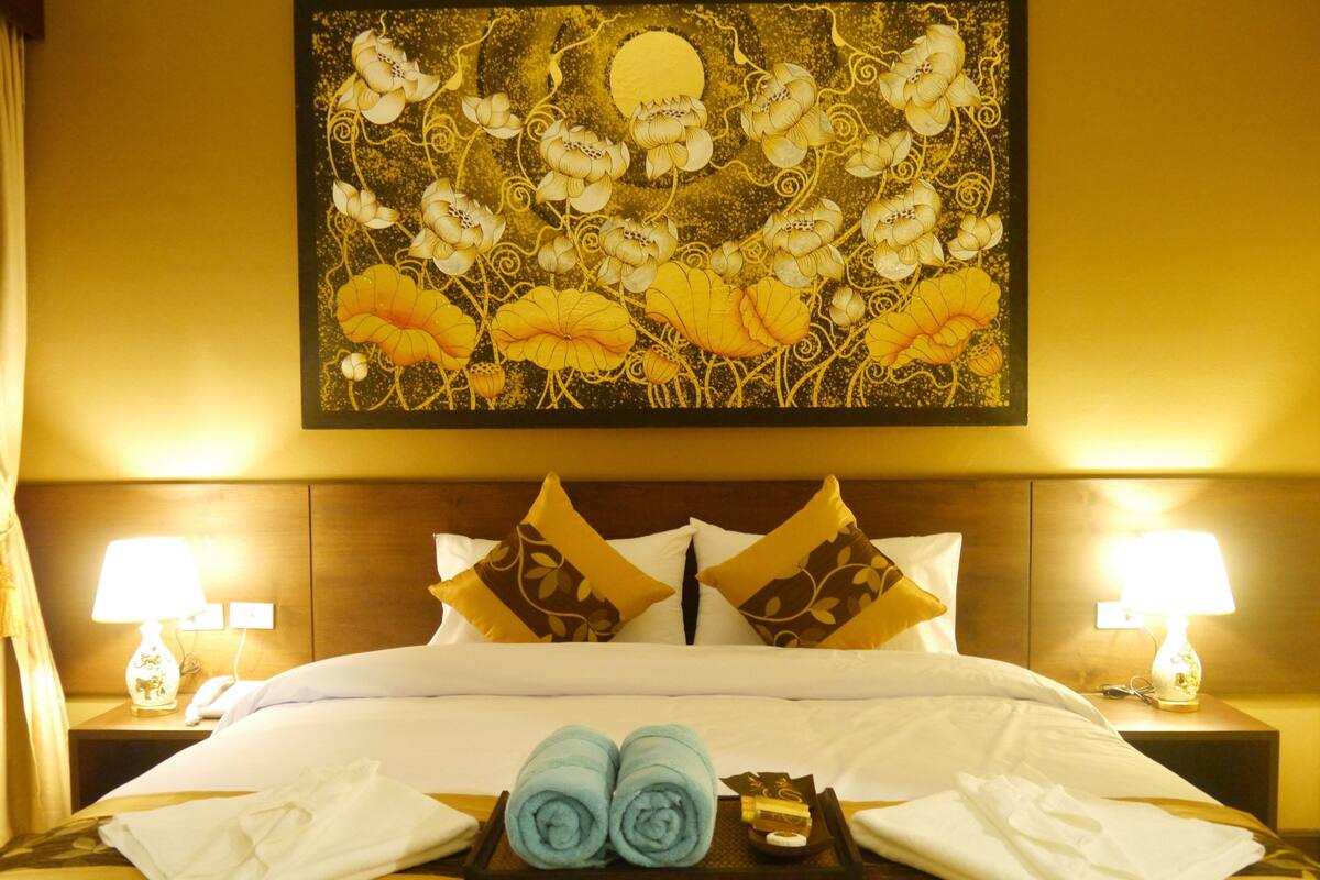Habitación de hotel con un cuadro floral grande en la pared, una cama bien hecha con ropa blanca, dos toallas enrolladas y una bandeja con artículos de aseo.
