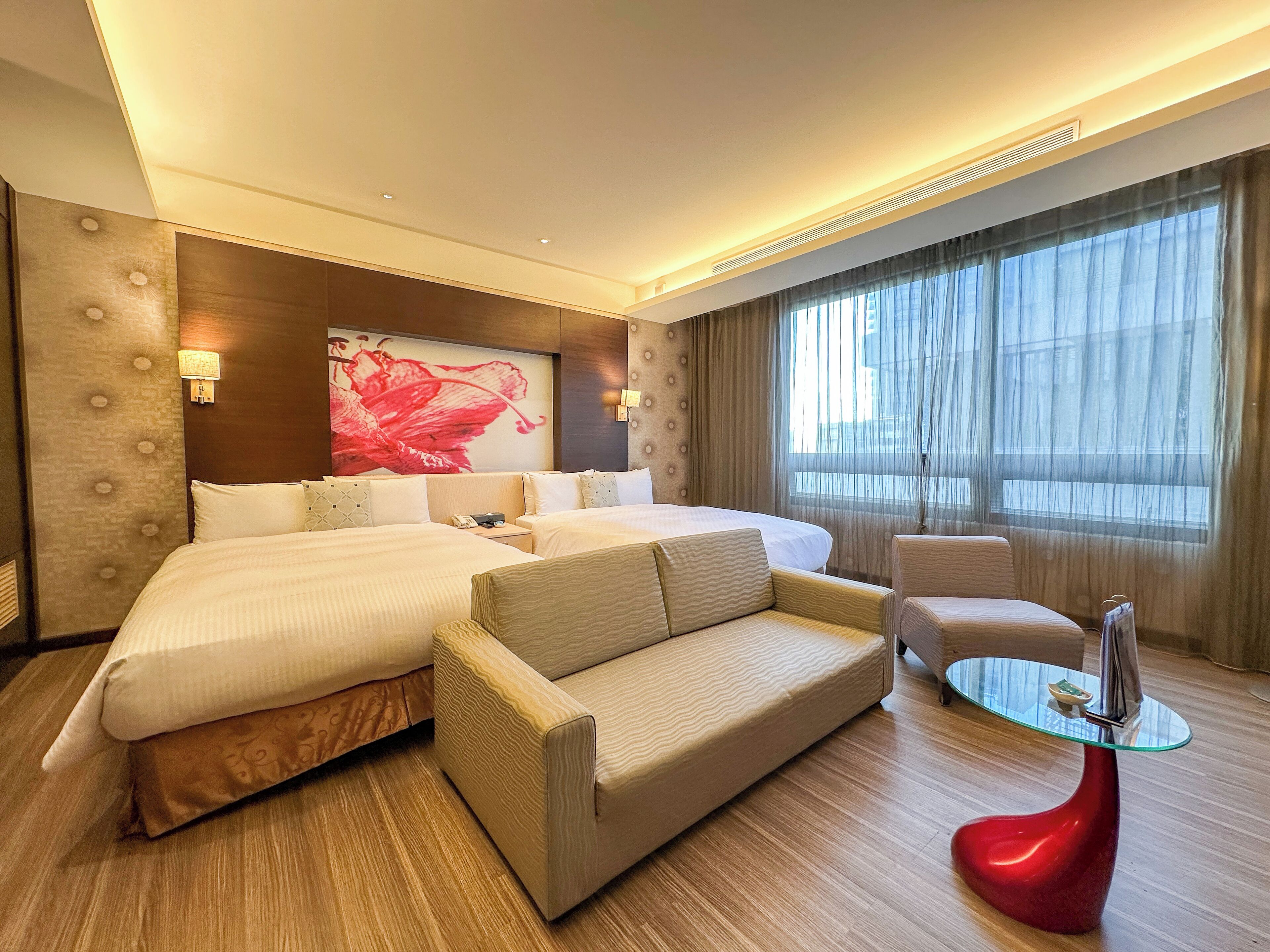 Habitación de hotel con una cama grande, un sofá, una mesita de vidrio y un ventanal con cortinas.