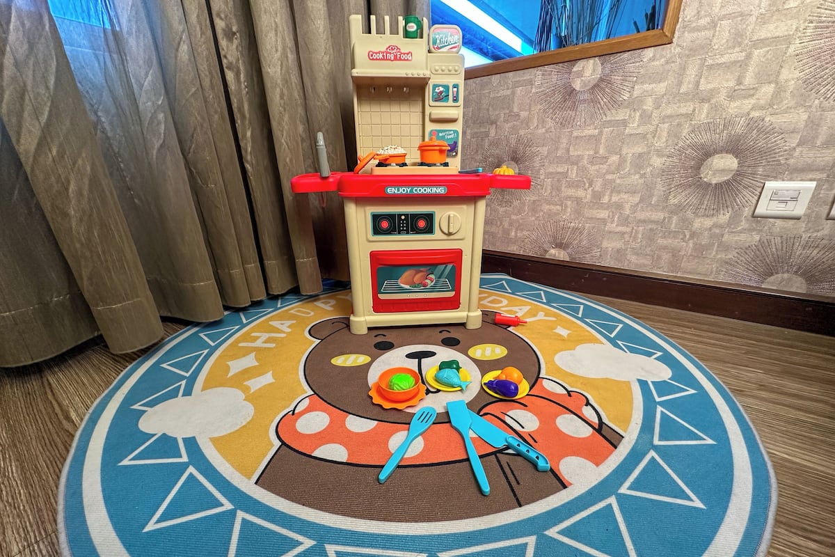 A play kitchen with a bear-themed rug, a window with curtains, and a brick-patterned wall.