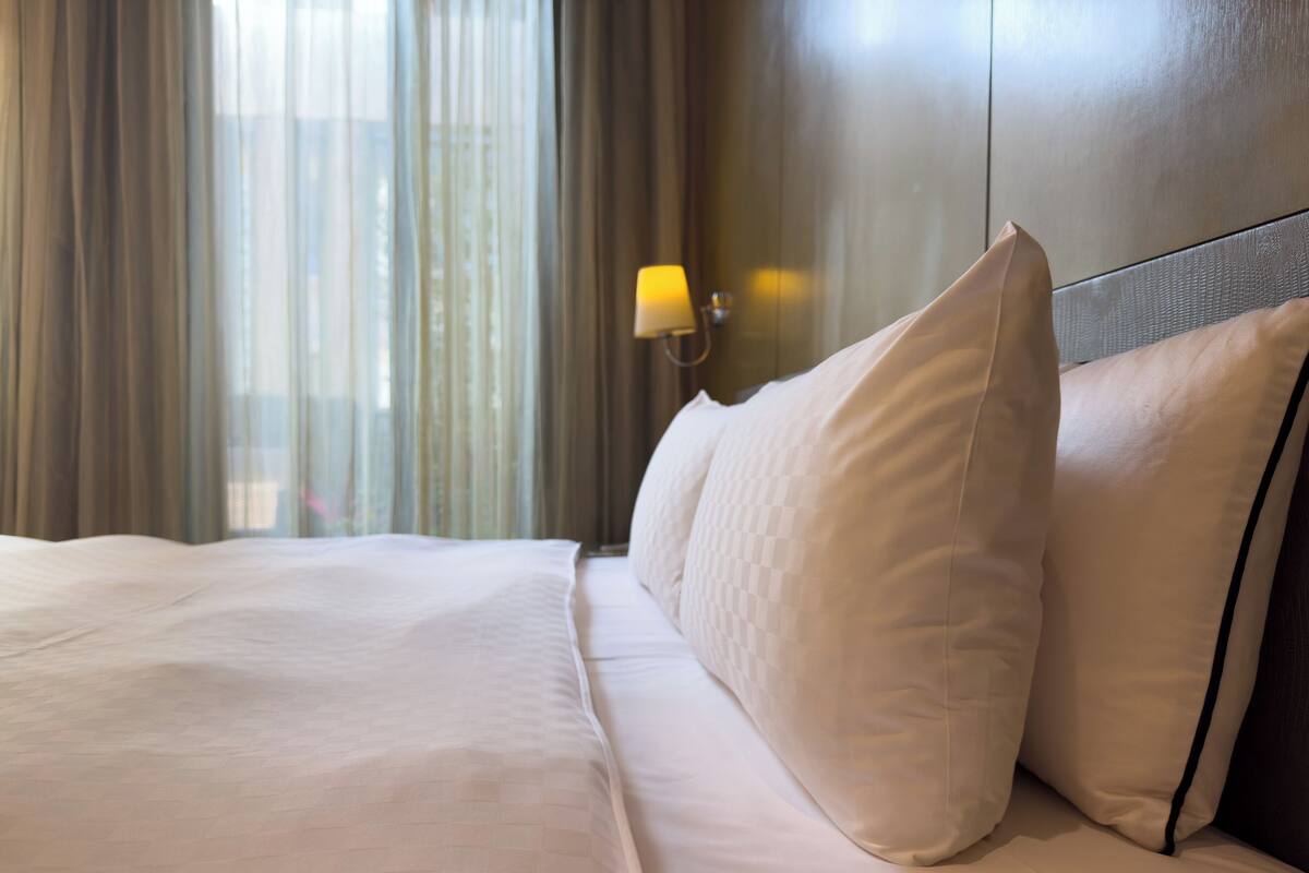 A hotel room with a bed, a nightstand with a lamp, and a view of the city through sheer curtains.