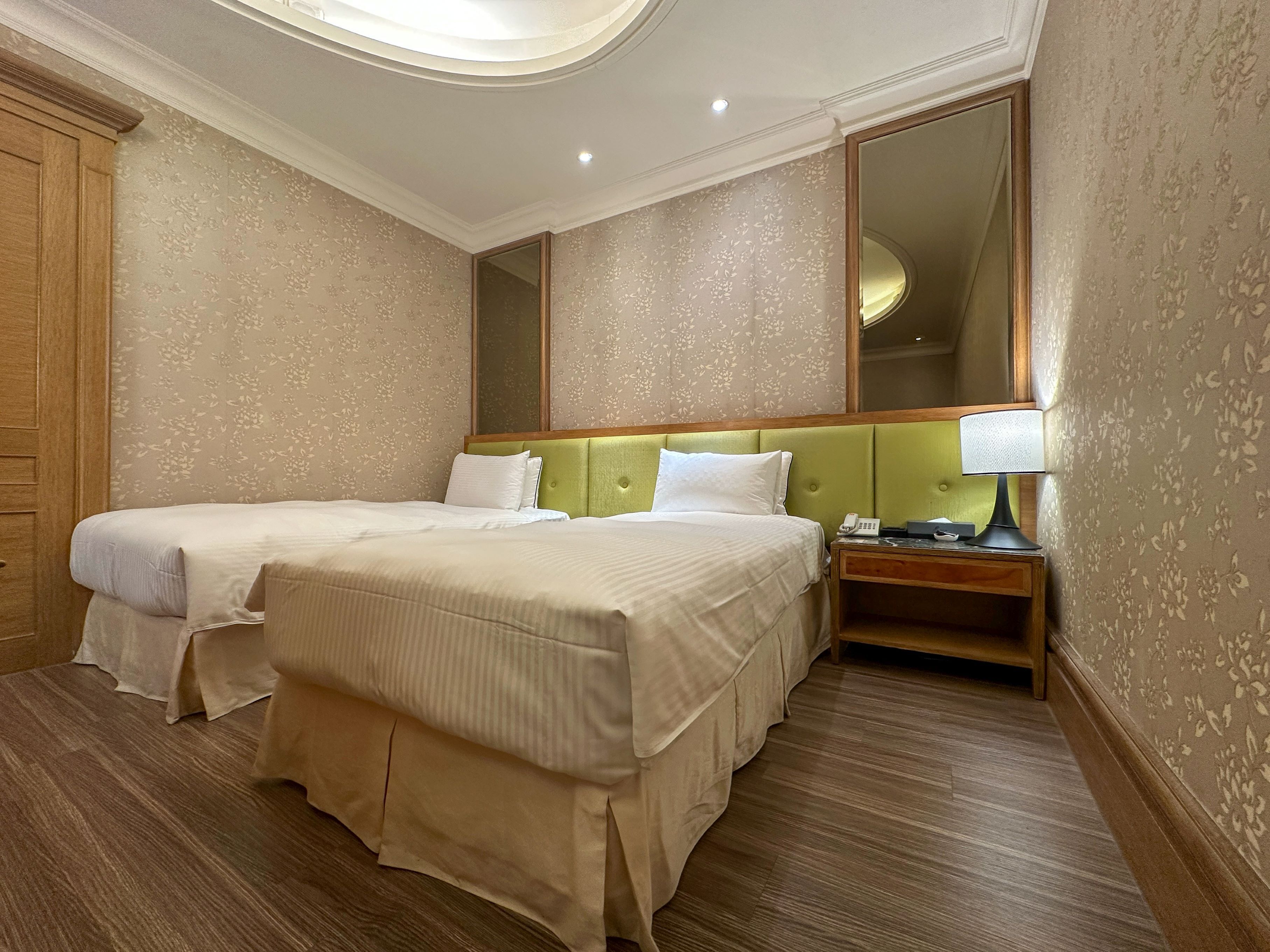 A hotel room with two beds, a nightstand with a lamp, and a large mirror on the wall.