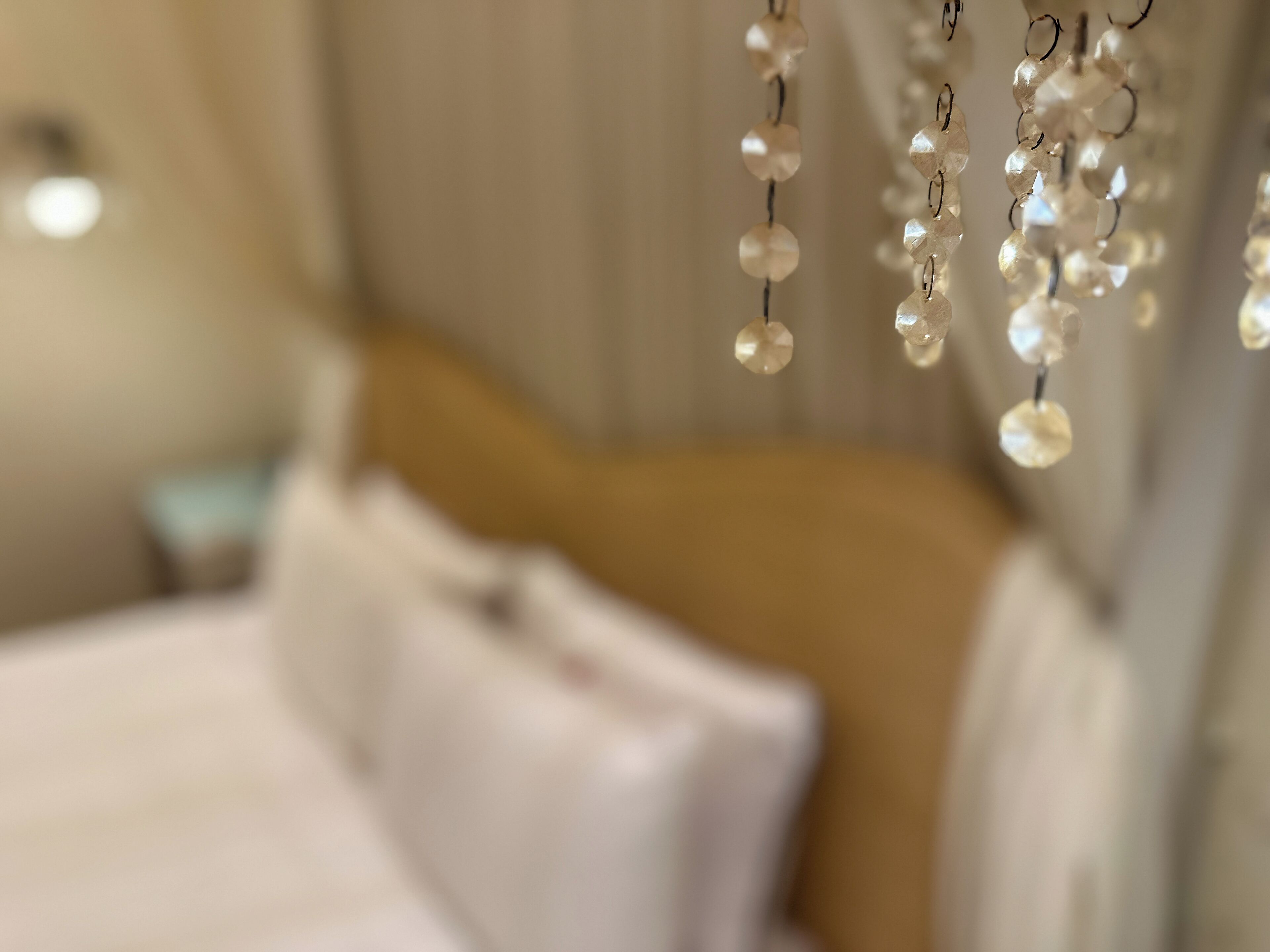 A bed with white linens and a headboard, a hanging light fixture with clear beads, and a blurred background suggesting a room with a window.
