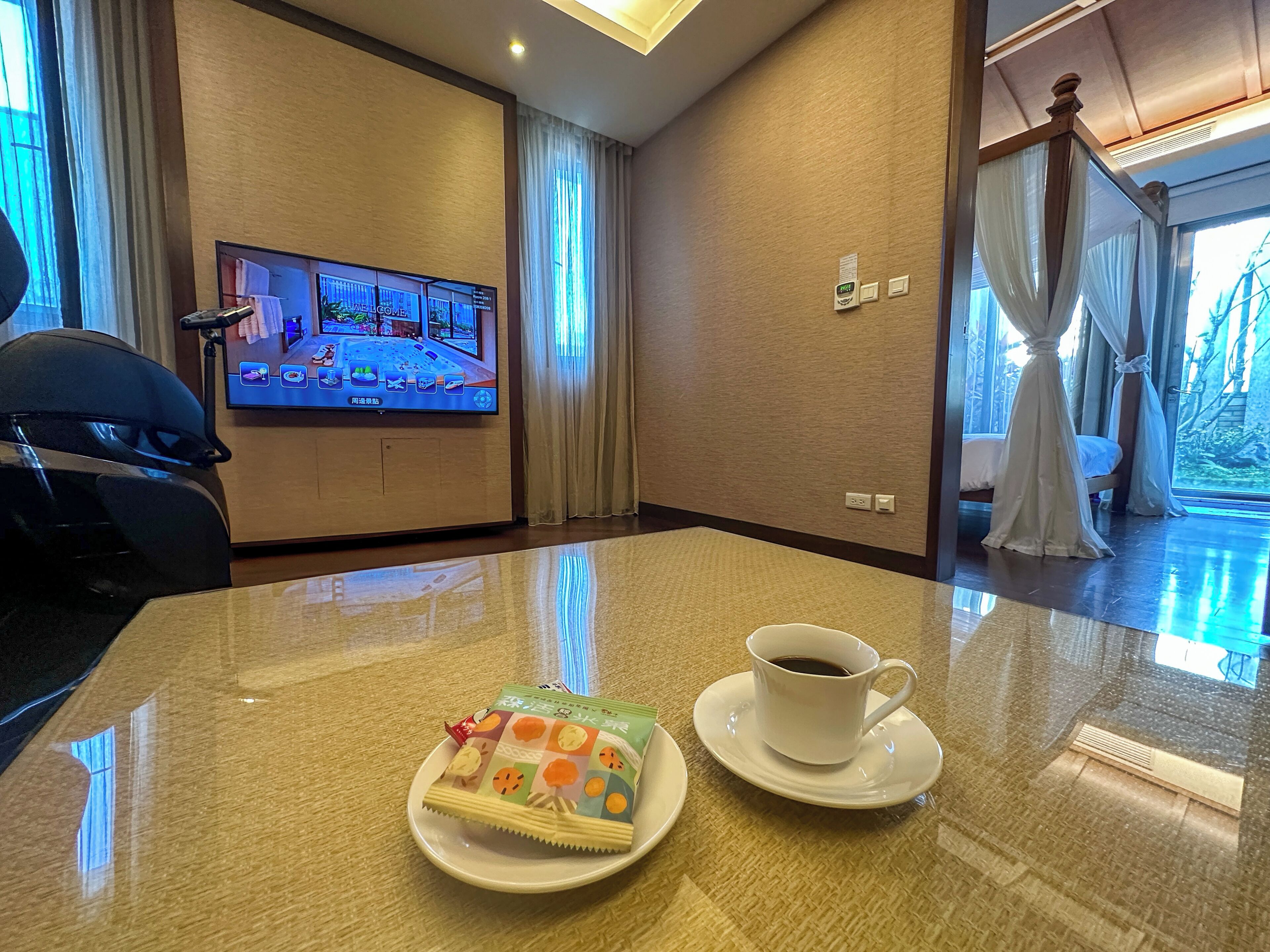 A hotel room with a large flat-screen TV on the wall, a dining table with a cup of coffee and a plate of snacks, and a bed with curtains.