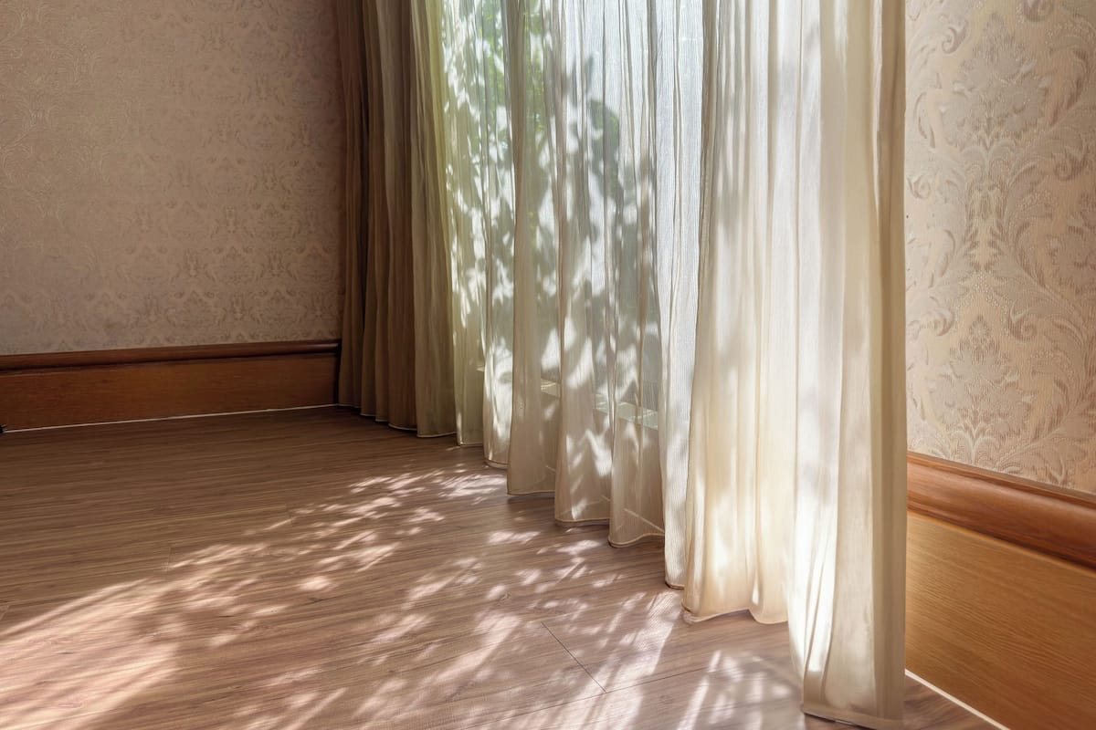 A room with wooden flooring, a patterned wallpaper, and sheer curtains allowing sunlight to filter through.