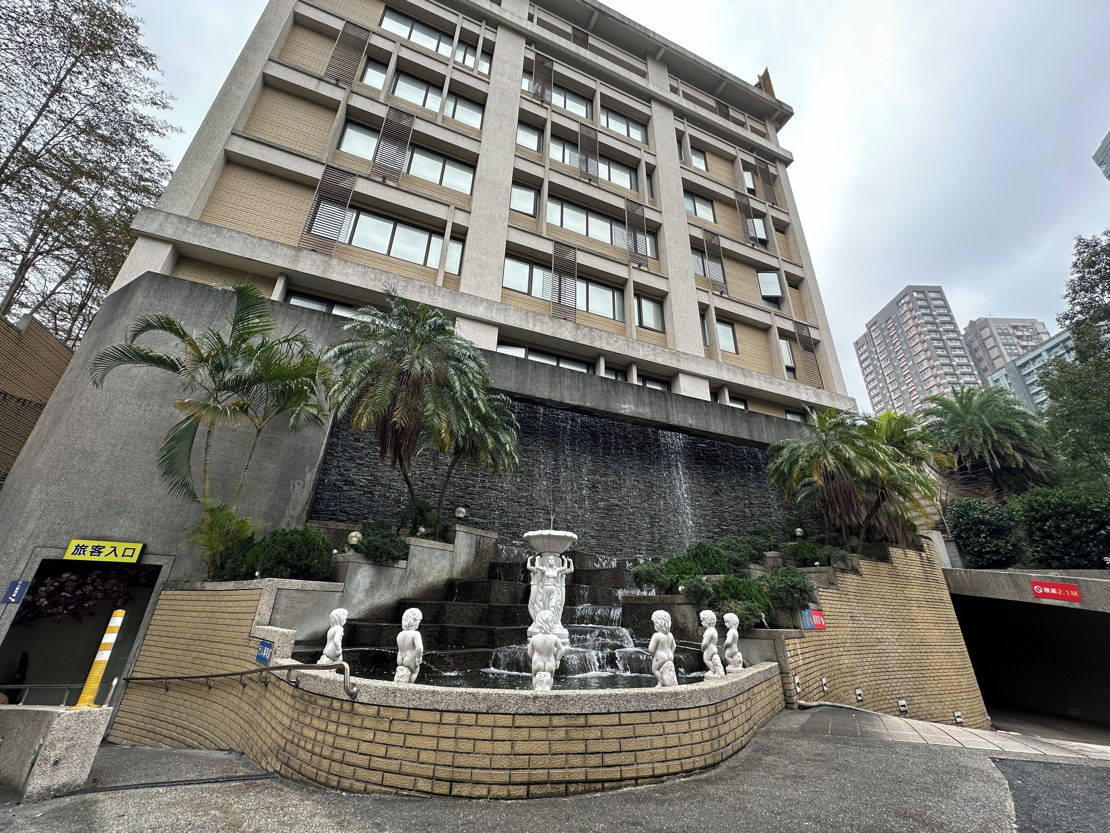 A modern building with a central water feature and statues, surrounded by palm trees and a stone wall.