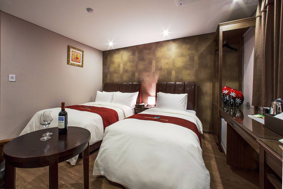 Habitación de hotel con dos camas, una mesita con una botella de vino, un espejo y un adorno rojo en la cómoda.
