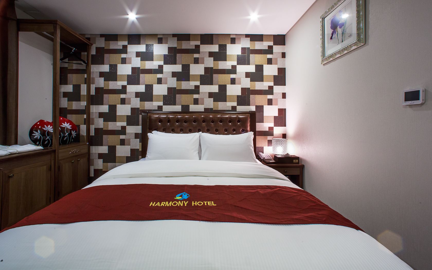A hotel room with a bed, a wooden wardrobe, a wall with a geometric pattern, a bedside table with a lamp, and a framed picture on the wall.