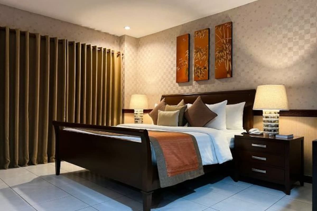 A modern hotel room with a large bed, two bedside tables with lamps, a nightstand, and a view of the city.