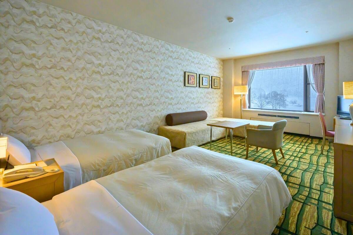 A hotel room with two beds, a small table, a sofa, a TV, a lamp, and a window with a view.