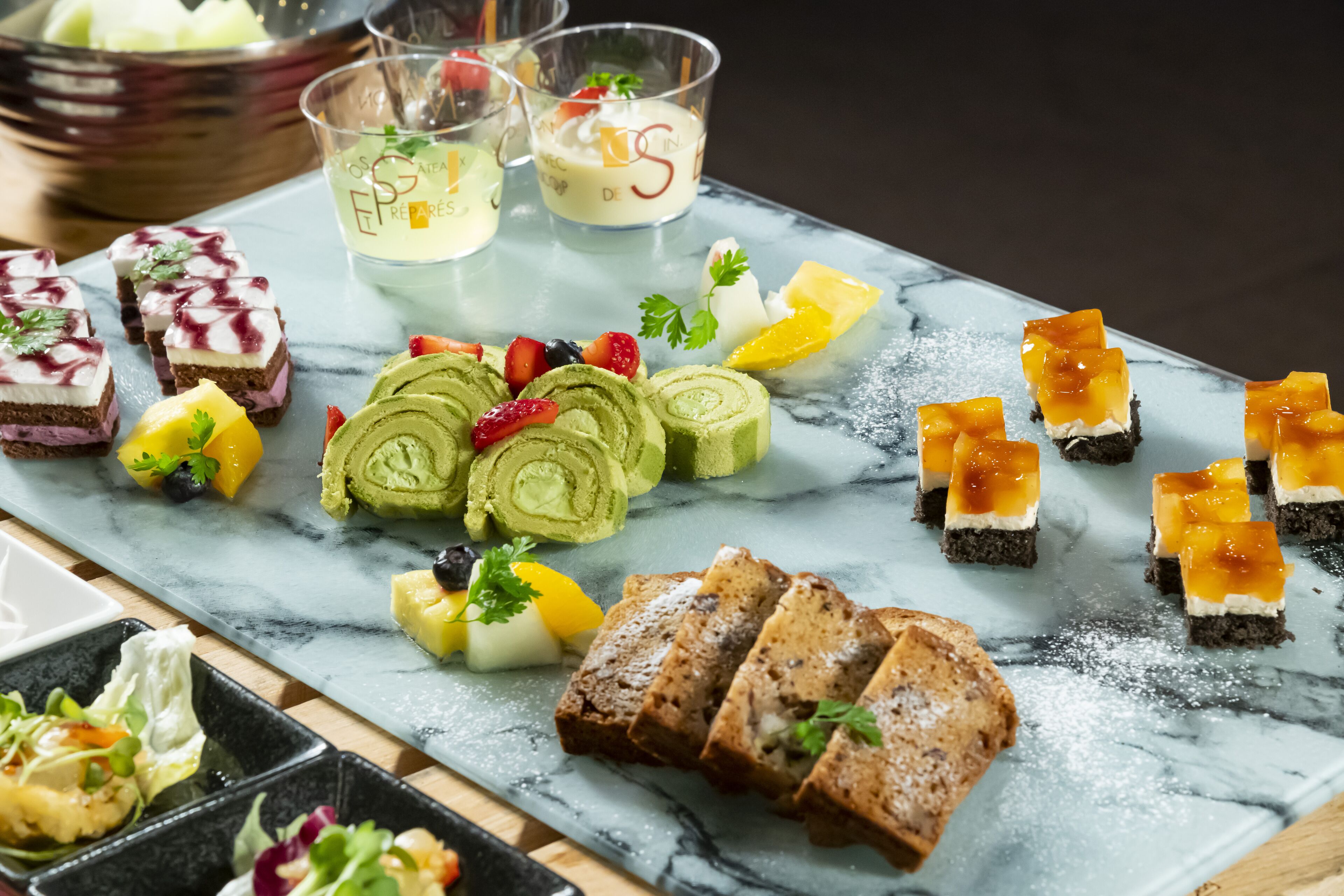 Una mesa con cubierta de mármol y diversos postres y pequeños platos, incluyendo postres enrollados de color verde, rodajas de fruta y pequeños recipientes de vidrio con crema.