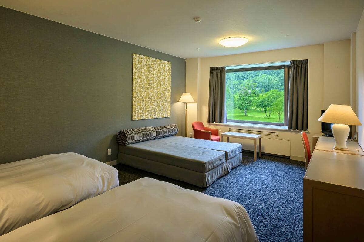A hotel room with two beds, a desk, a chair, and a window with a view of greenery.