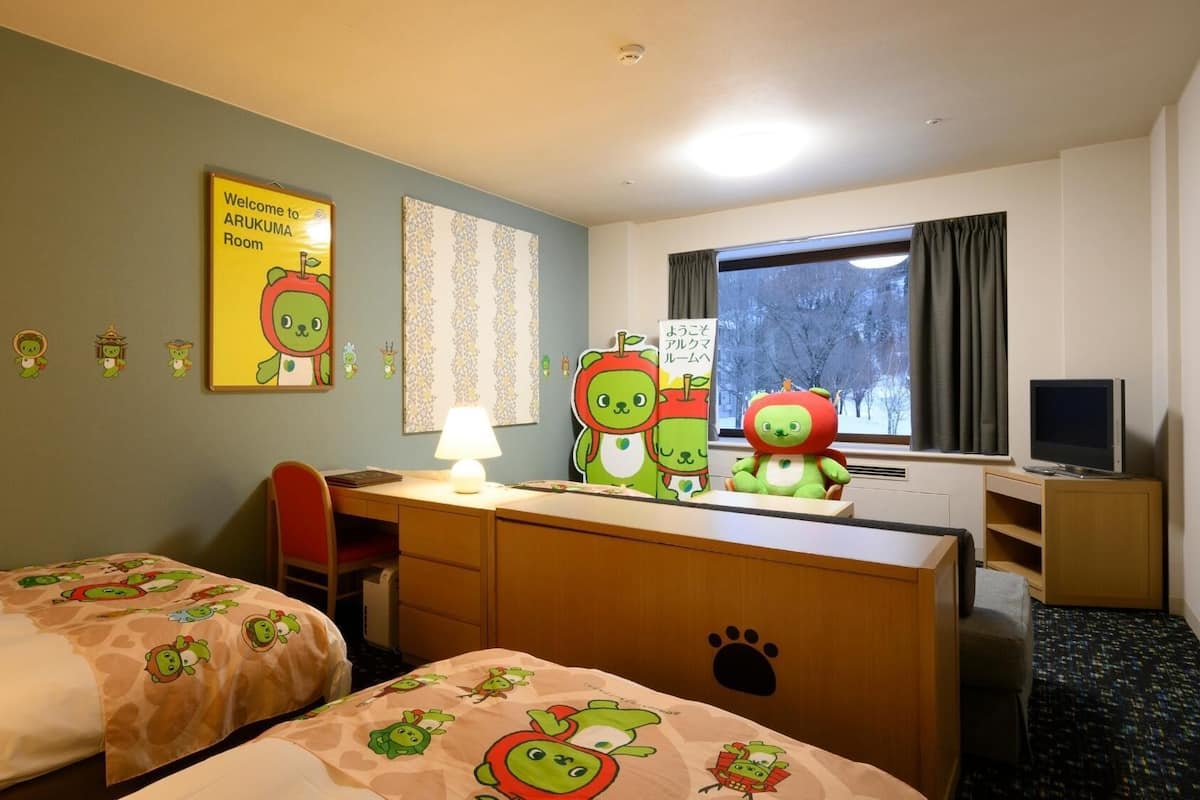 Habitación de hotel con dos camas, un escritorio con lámpara, televisión y ventana con vista a árboles.