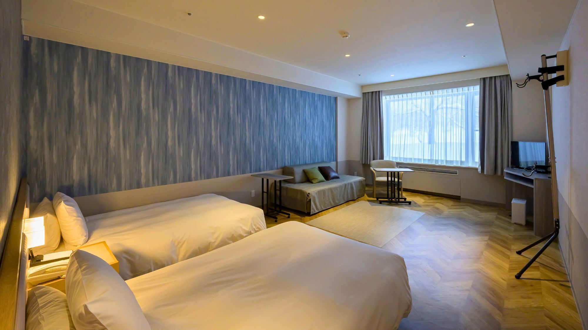 A hotel room with two beds, a sofa, a desk with a TV, and a large window with curtains.