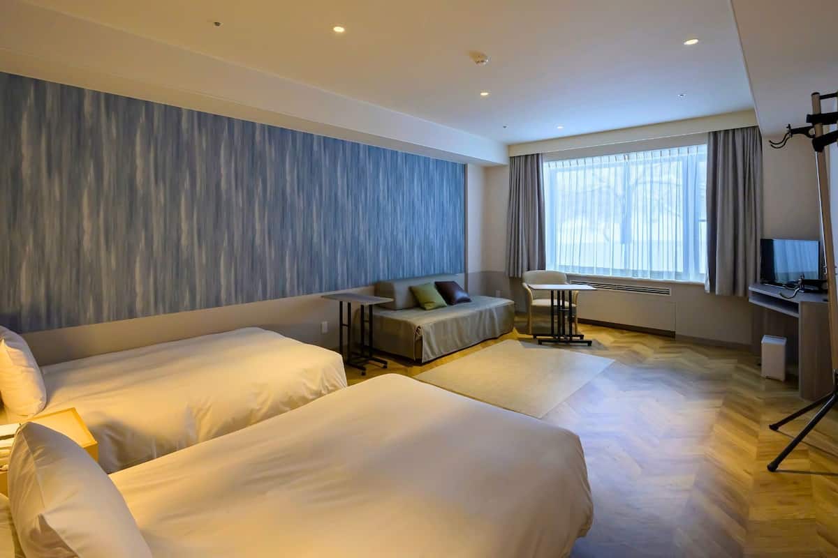 Habitación de hotel con dos camas, un sofá, un escritorio con televisor y un amplio ventanal con cortinas.