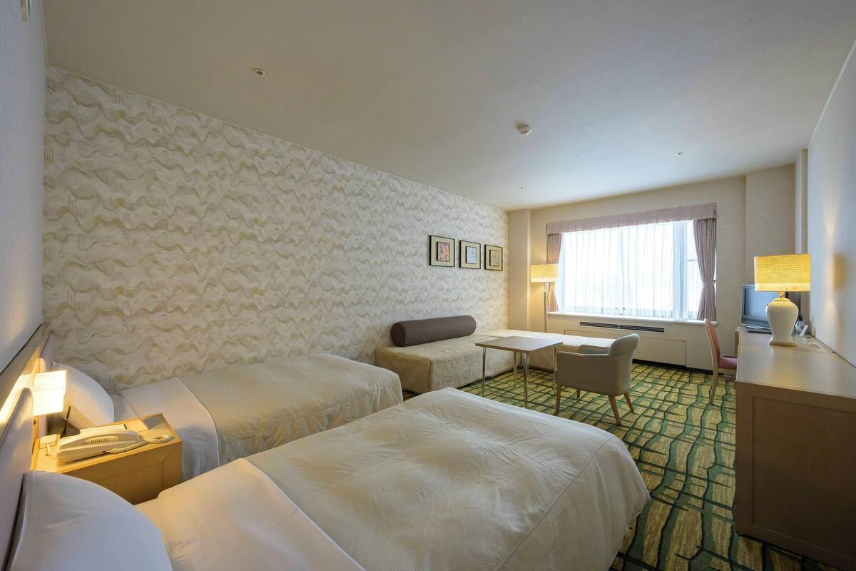 A hotel room with two beds, a small table, a chair, a desk, and a window with curtains.