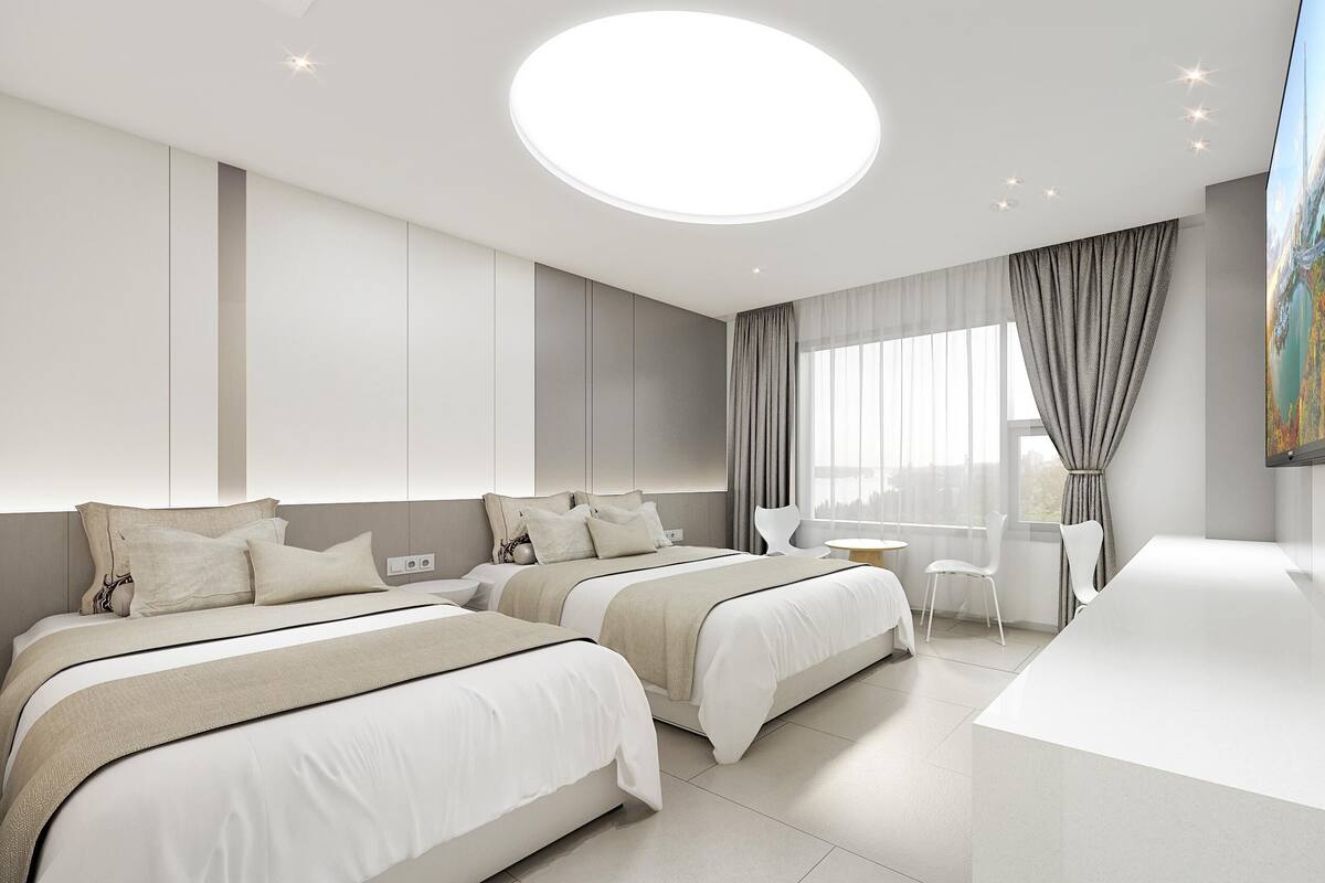 A modern hotel room with two beds, a large window with curtains, a flat-screen TV, and a round ceiling light.