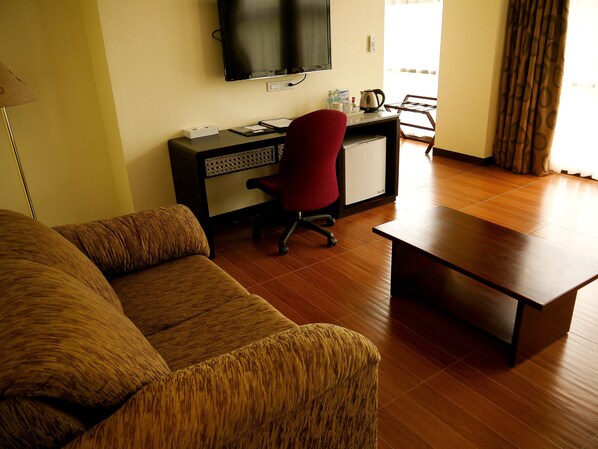 Un salon avec un canapé, un fauteuil de bureau rouge, un petit bureau avec un ordinateur et un téléviseur fixé au mur.