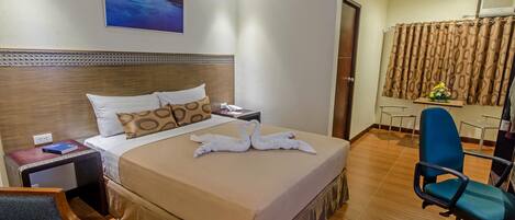 A hotel room with a large bed, a wooden headboard, a nightstand, a desk with a chair, a blue office chair, and a picture on the wall.
