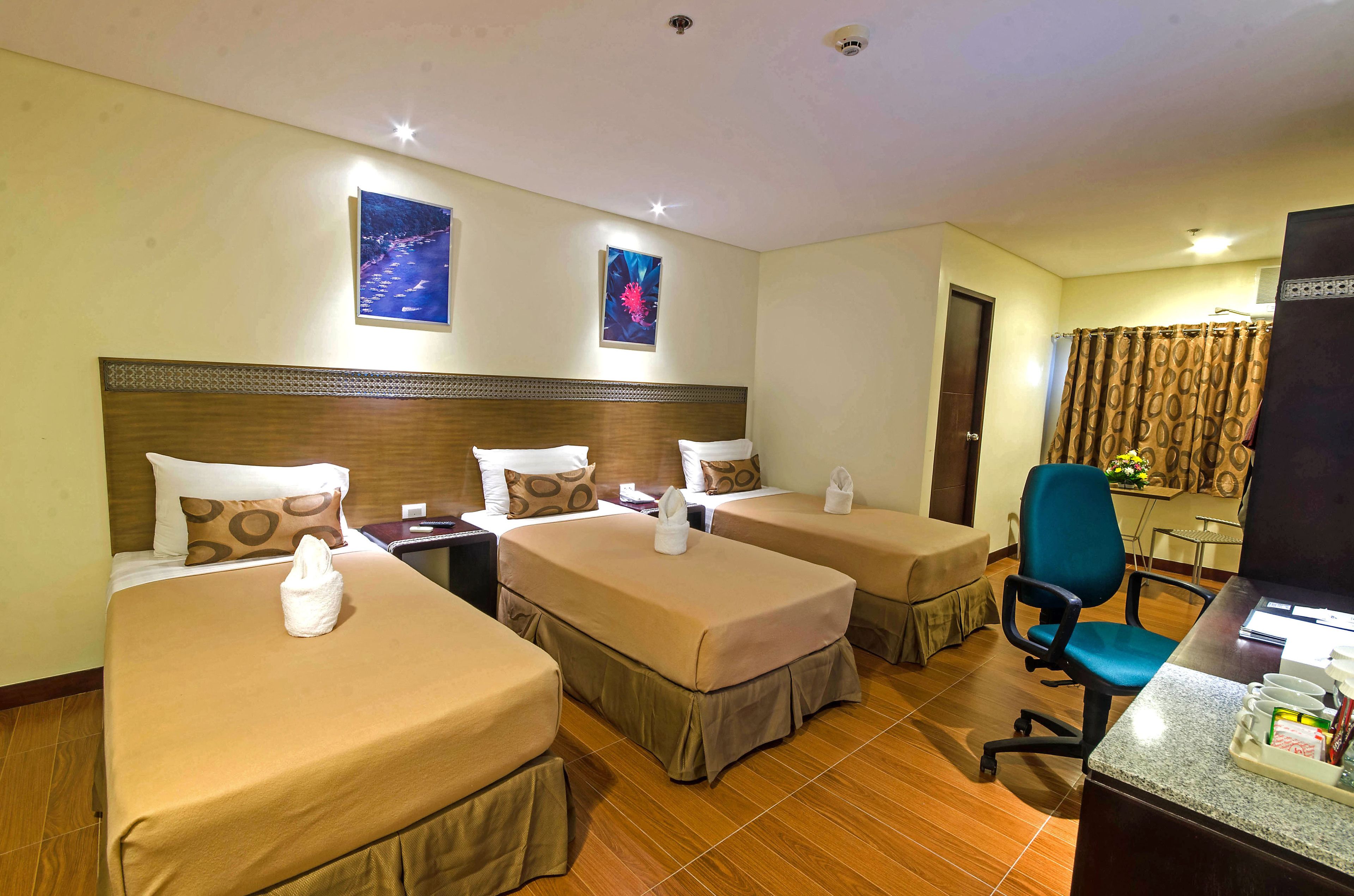 Une chambre d’hôtel avec trois lits simples, un bureau avec une chaise et deux téléviseurs fixés au mur.