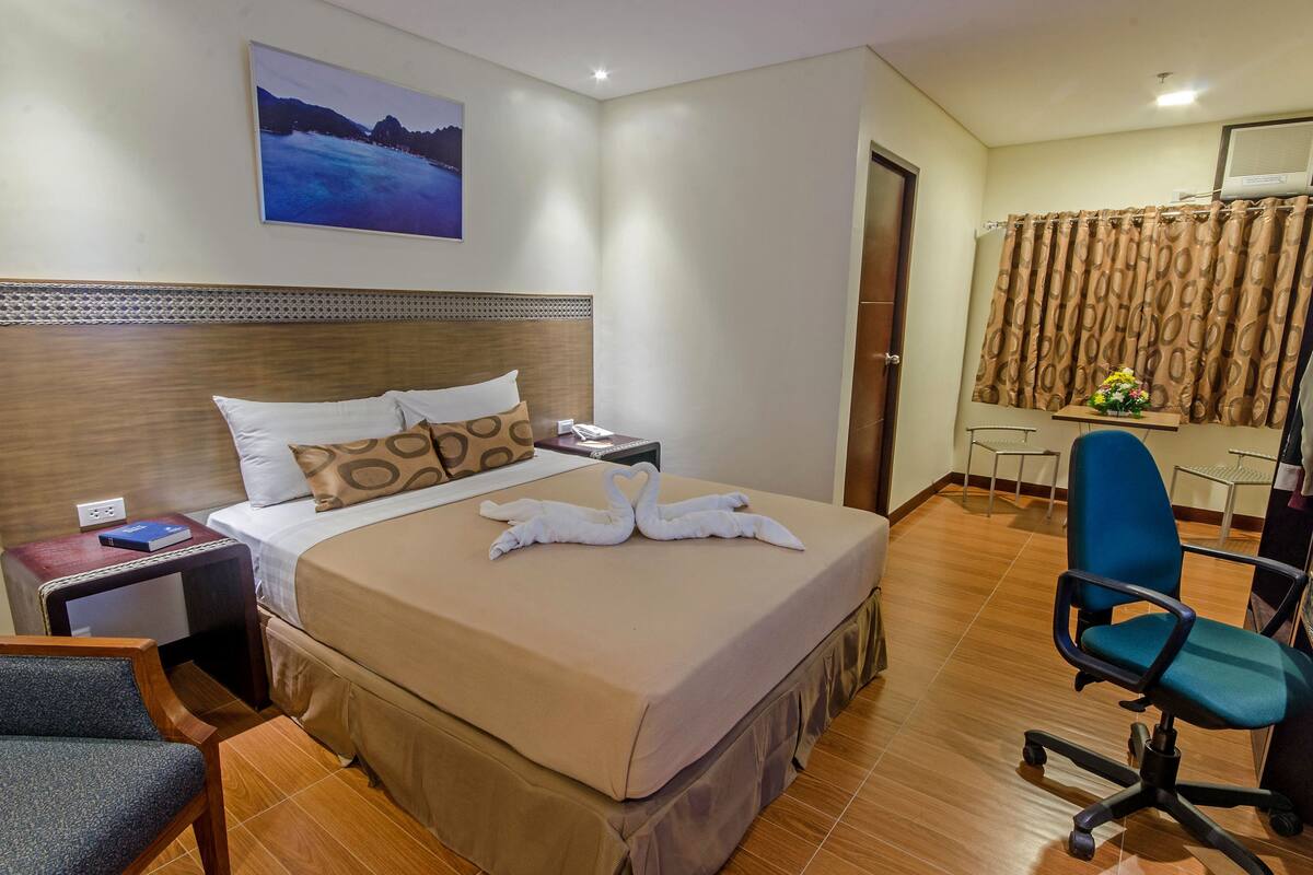 Ein Hotelzimmer mit einem großen Bett, einem Schreibtisch, einem Sessel, einem Bild an der Wand und einem Vorhang.