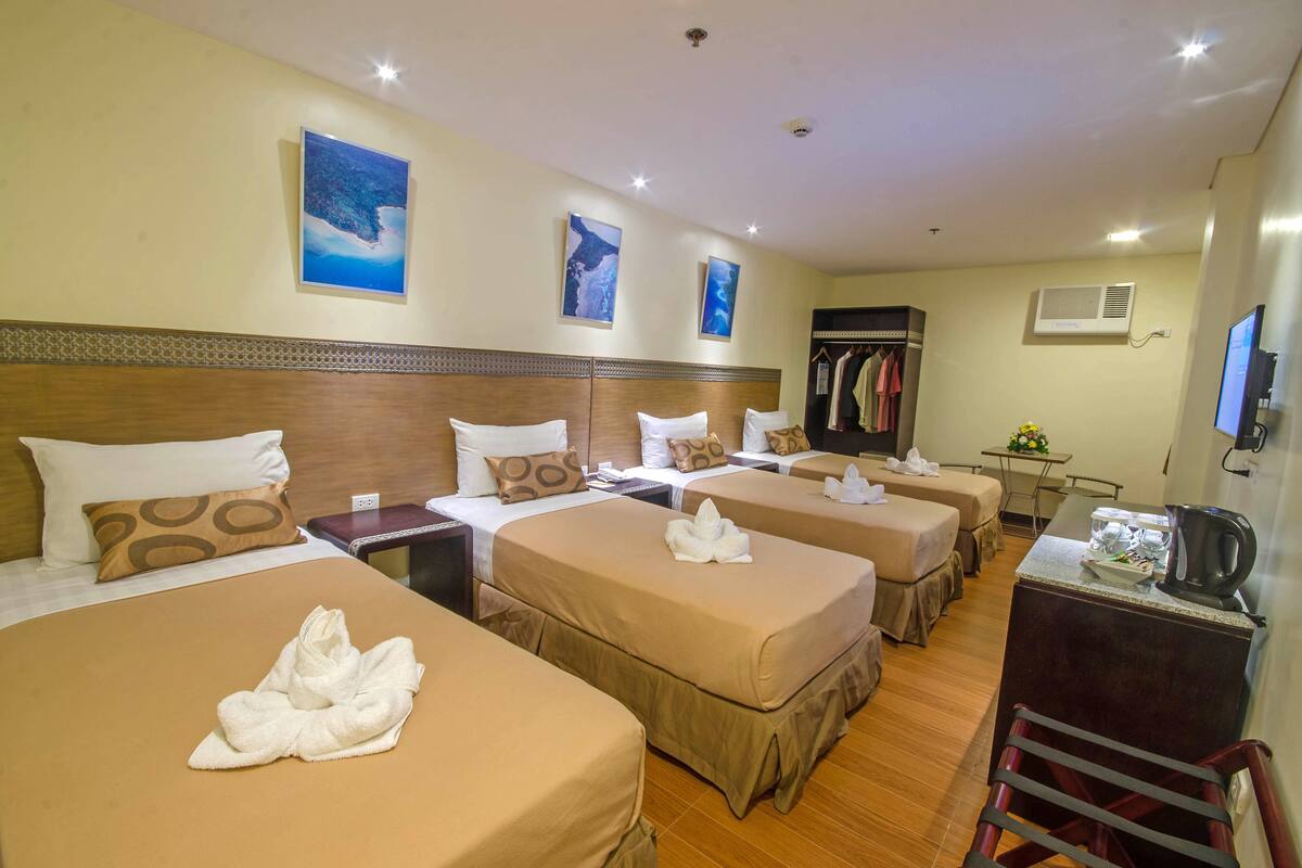 Ein Hotelzimmer mit vier Betten, jeweils mit einer braunen Tagesdecke und weißen Kissen. An der Wand befinden sich Wandklimaanlagen und ein Fernseher.