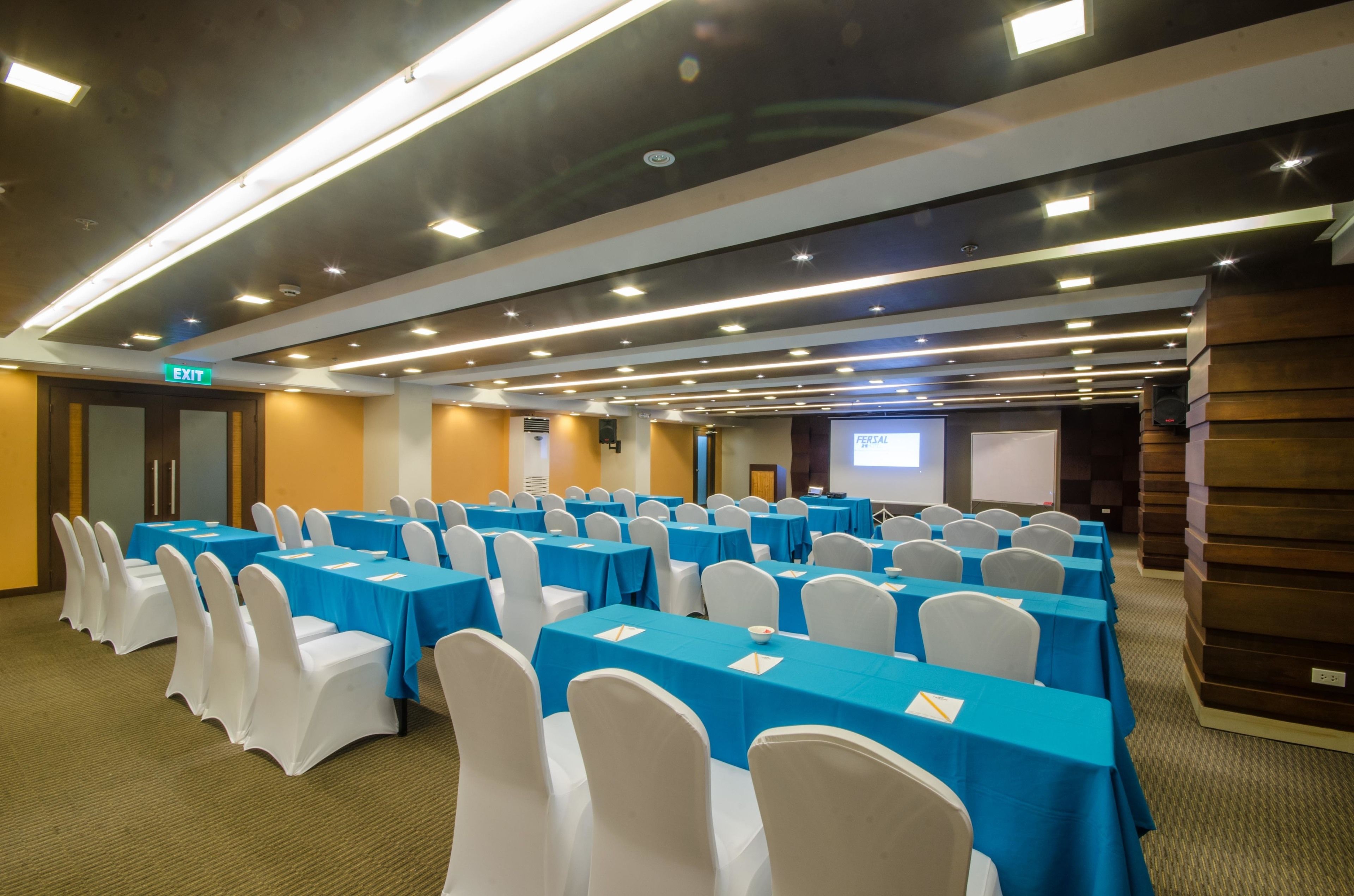 A conference room with rows of tables and chairs, a projector screen, and an exit sign.