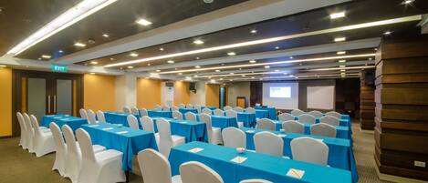 A conference room with rows of tables and chairs, a projector screen, and an exit sign.