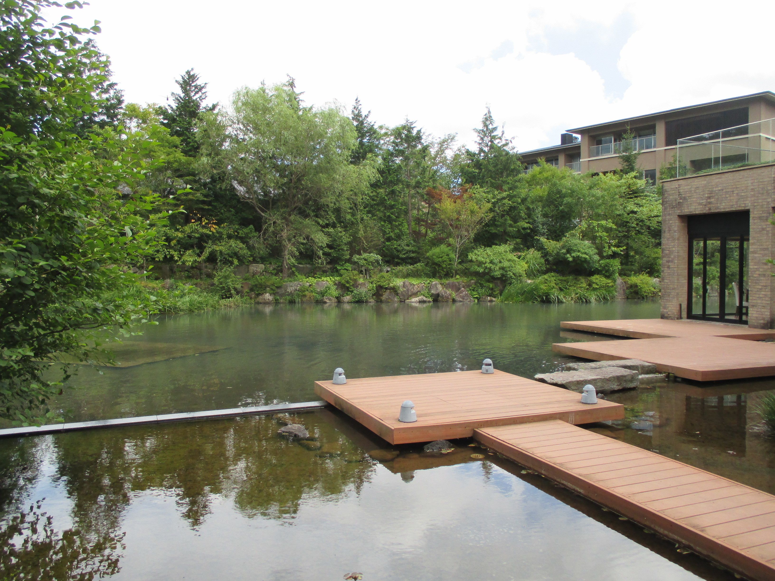 A tranquil outdoor area with a wooden deck over water, surrounded by lush greenery and a modern building with large windows.