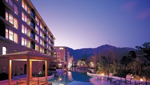Un hotel con piscina, mobiliario exterior y montañas al fondo.