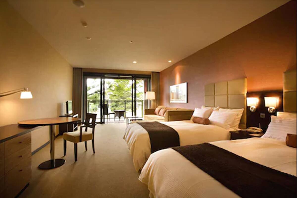 A hotel room with two beds, a desk, a chair, and a window with a view of greenery.