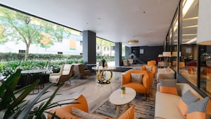 Un área de descanso moderna con asientos anaranjados, una mesa redonda y ventanas amplias con vistas a un jardín.
