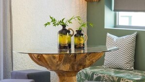 A modern room with a glass-top table, two grey ottomans, a green patterned sofa, and a wall-mounted lamp. - Sunshine Hotel (Malia)