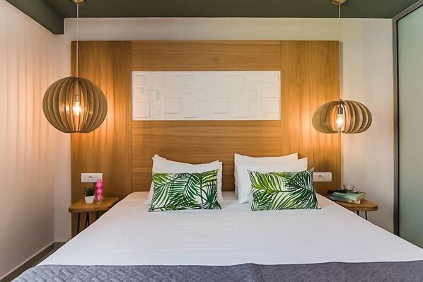 A hotel room with a large bed, two bedside tables, a wooden headboard, and two pendant lights. - Sunshine Hotel (Malia)