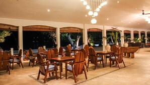 Un vaste espace extérieur aménagé pour les repas, avec des tables et des chaises en bois, de grands pots de plantes et une vue sur l’océan.
