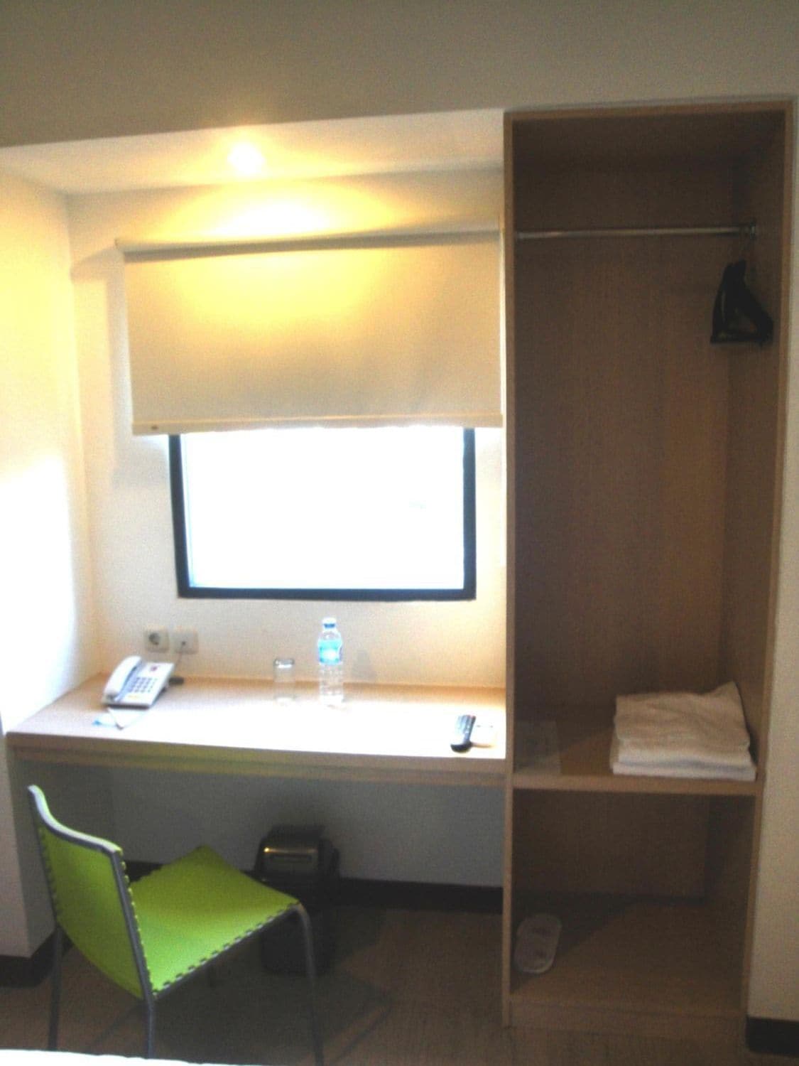 A small room with a desk, a chair, a window, a wardrobe, and a bedside table with a bottle of water and a phone.