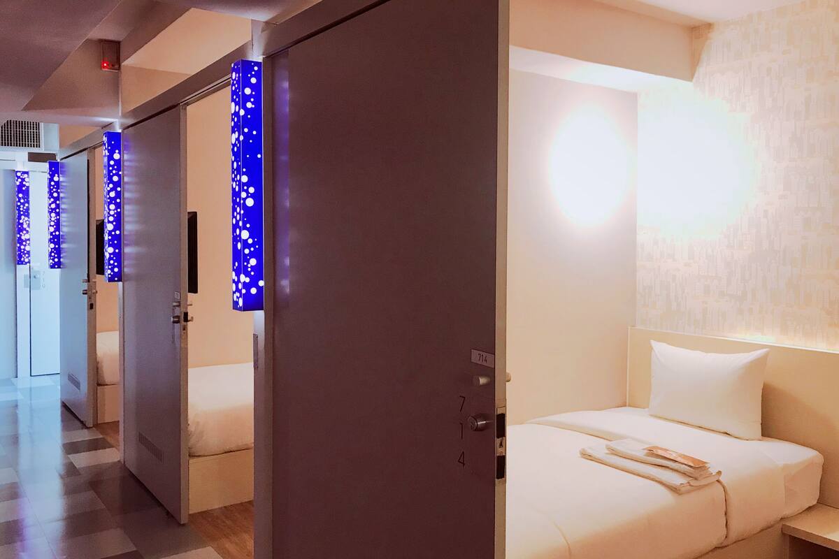 A hotel room with a bed, a nightstand, and a wall with a cityscape wallpaper. There are doors with blue lights and a check-in counter in the background.