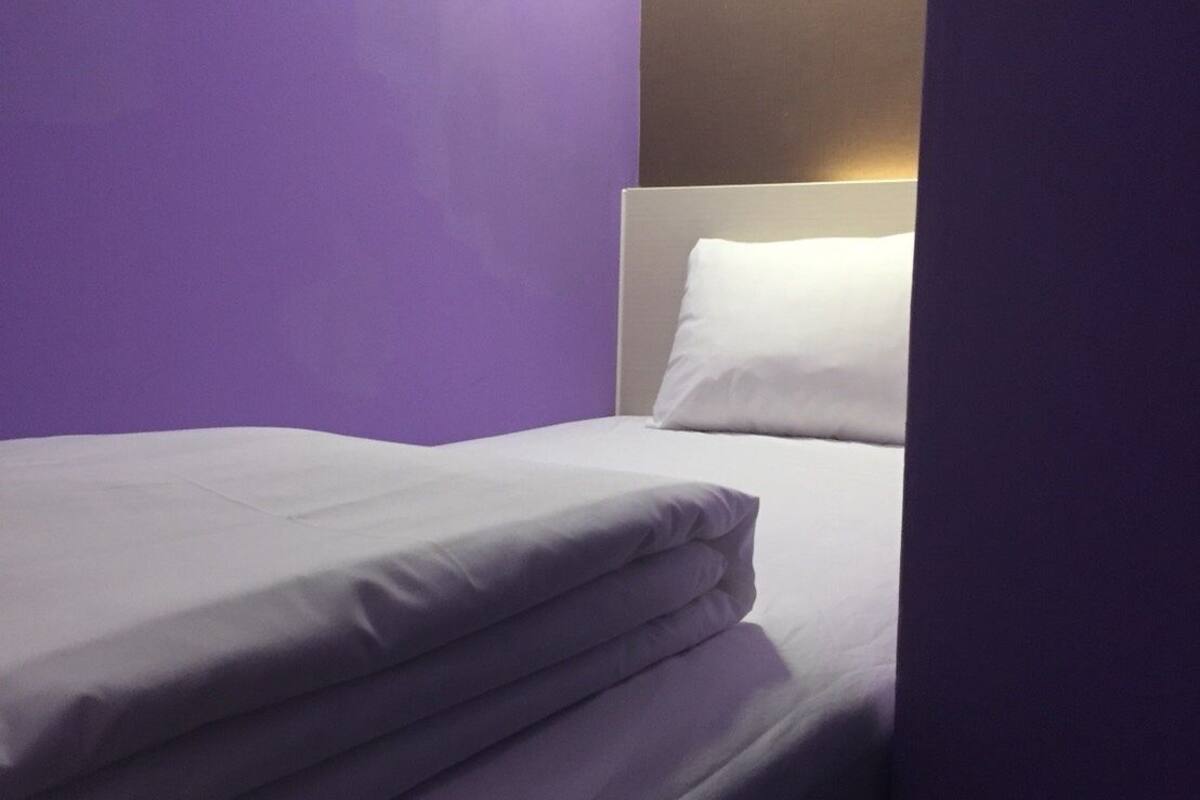 A single bed with white bedding in a room with purple walls and a light source above the bed.
