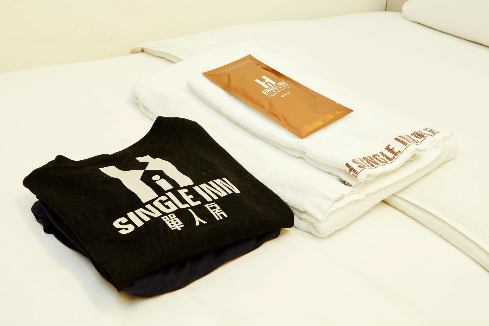 Un t-shirt noir, soigneusement plié, avec un texte blanc, et une serviette blanche également ornée d’un texte, sont posés sur un lit.