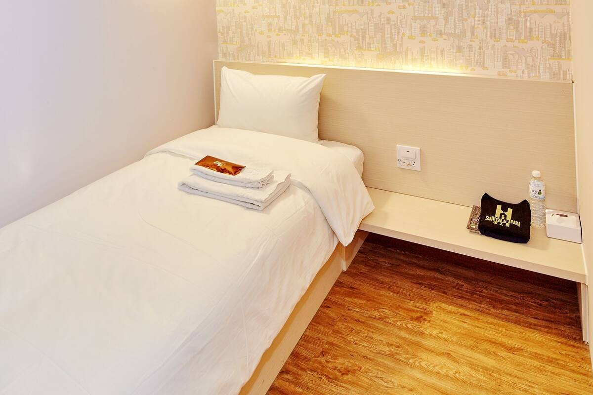 A single bed with white bedding, a small bedside table with a black bag and a bottle, and a patterned wallpaper on the wall.