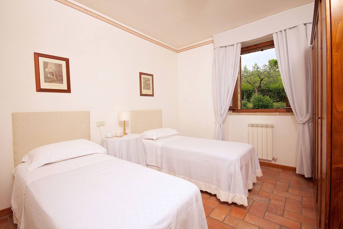 A hotel room with two single beds, a window with curtains, a radiator, and framed pictures on the wall.