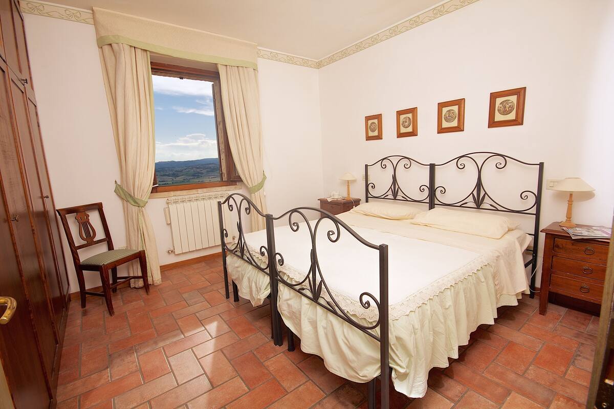 A bedroom with a bed, a chair, a window with curtains, a radiator, and a wall with framed pictures.