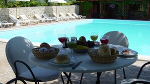 Area ristoro a bordo piscina con tavolo apparecchiato per due, con frutta, pane e bevande assortite.