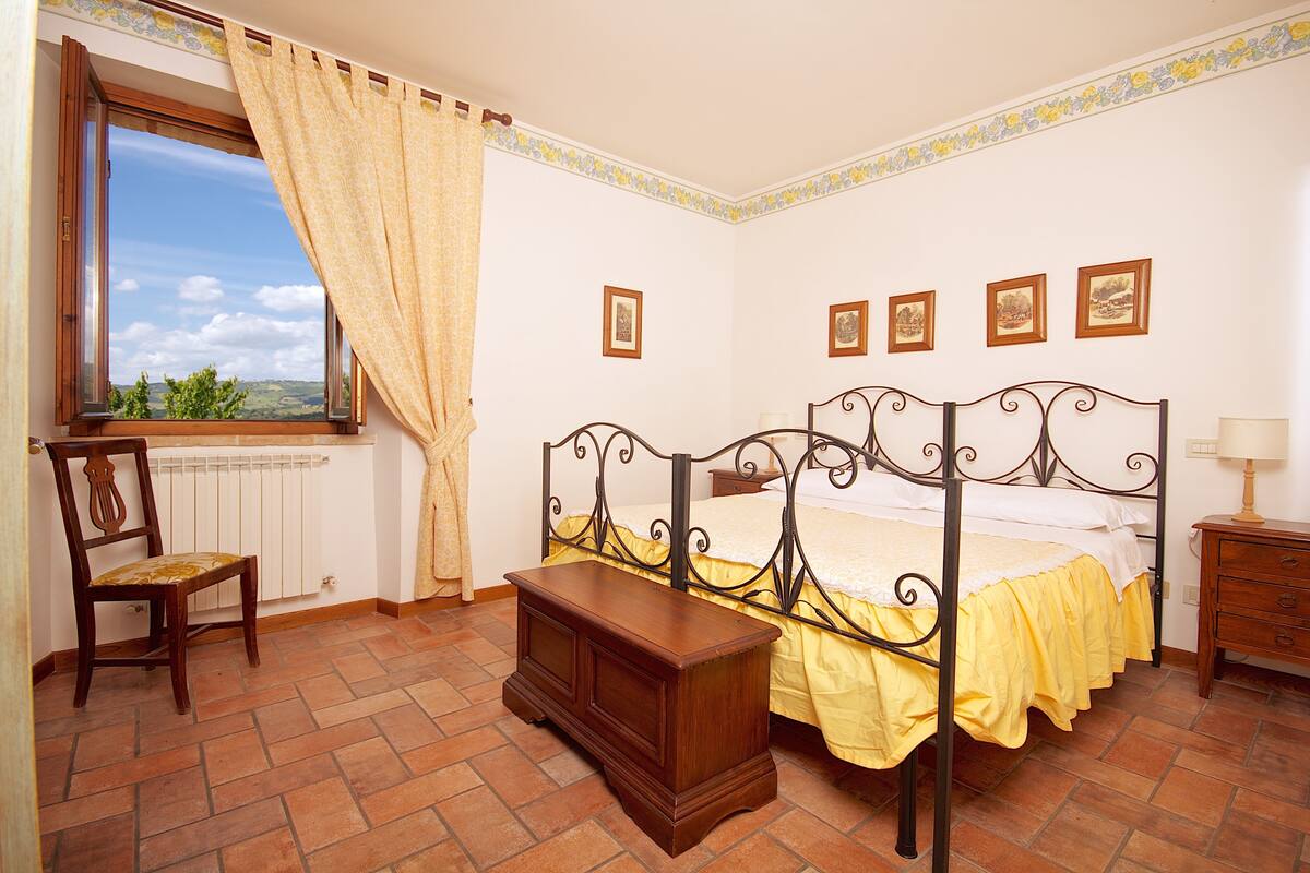A bedroom with a four-poster bed, a wooden nightstand, a chair, and a window with curtains.