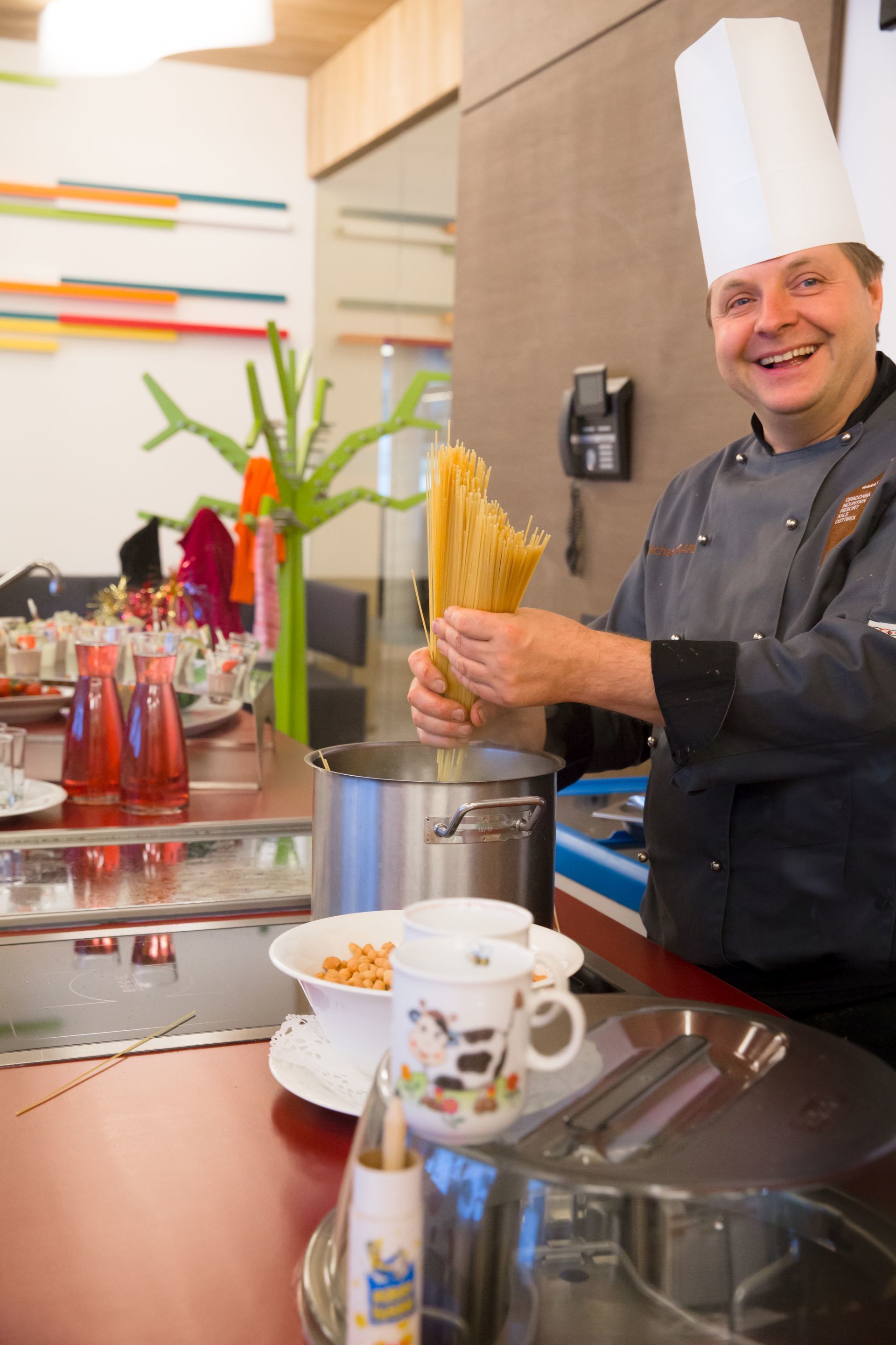 A chef in a professional kitchen holding uncooked pasta, with cooking utensils and a pot on the counter.