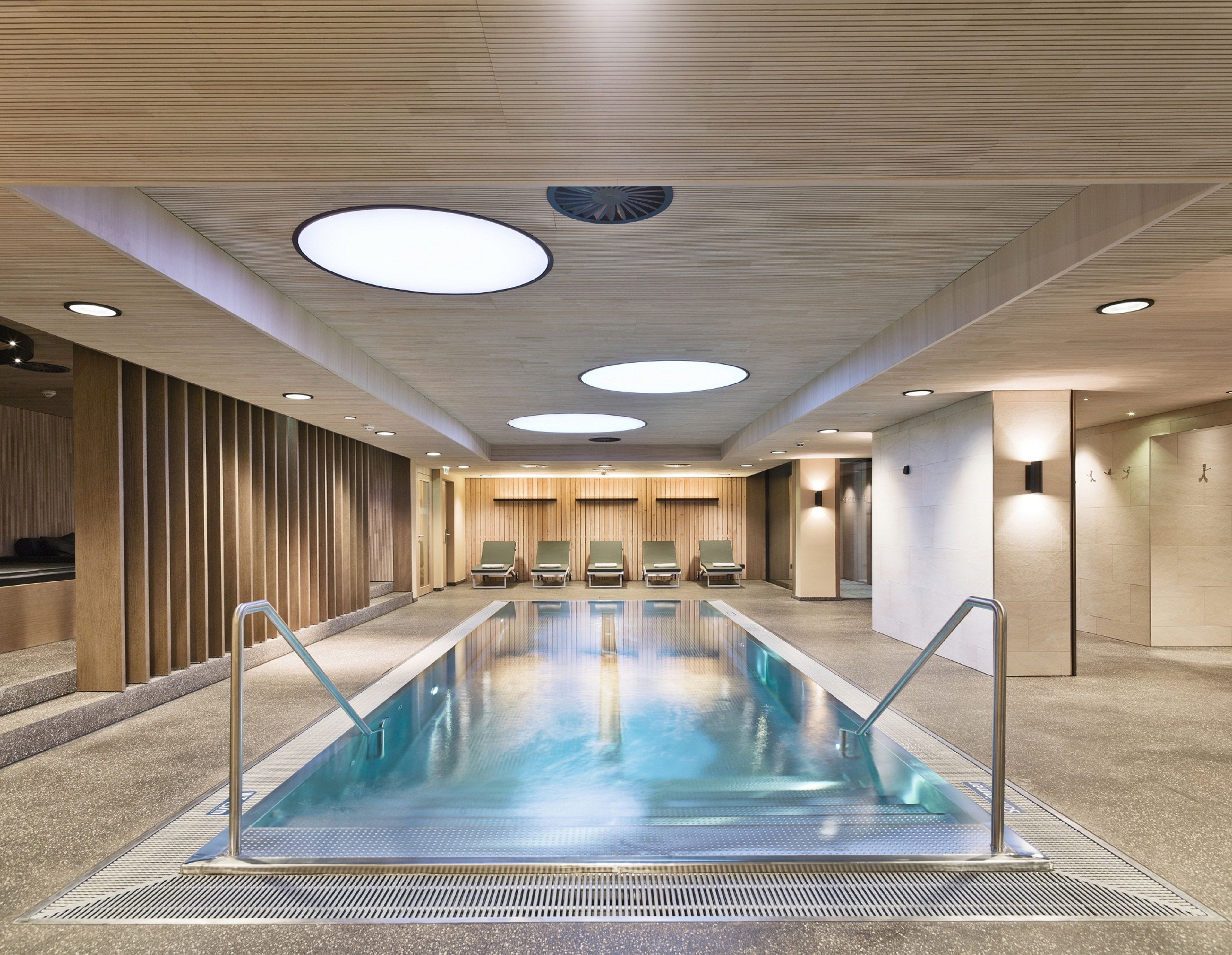 Une piscine intérieure moderne, avec une eau cristalline, entourée de chaises longues et agrémentée d’un mur en panneaux de bois.