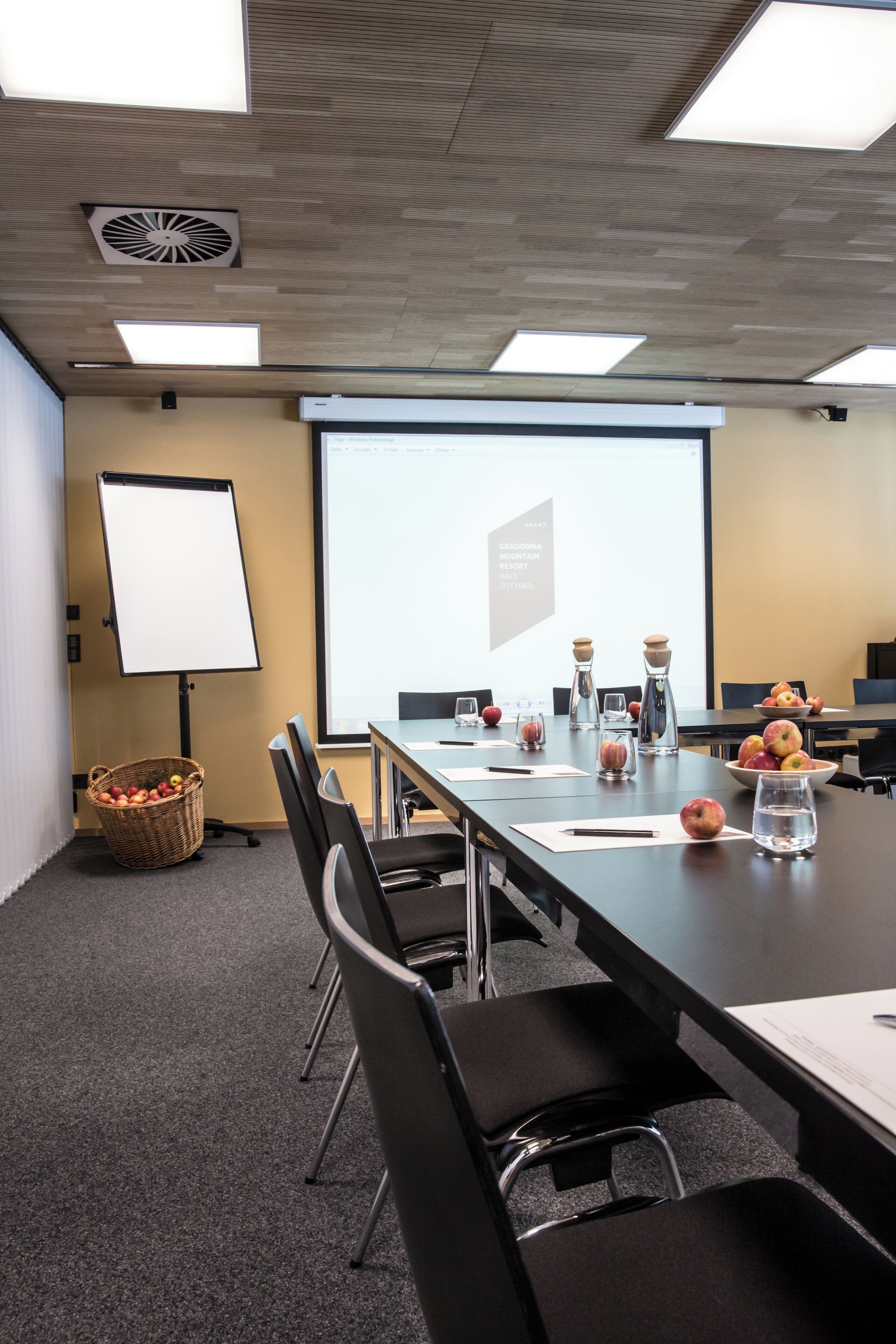 A conference room with a long table, chairs, a projector screen, a whiteboard, and a basket of fruit.