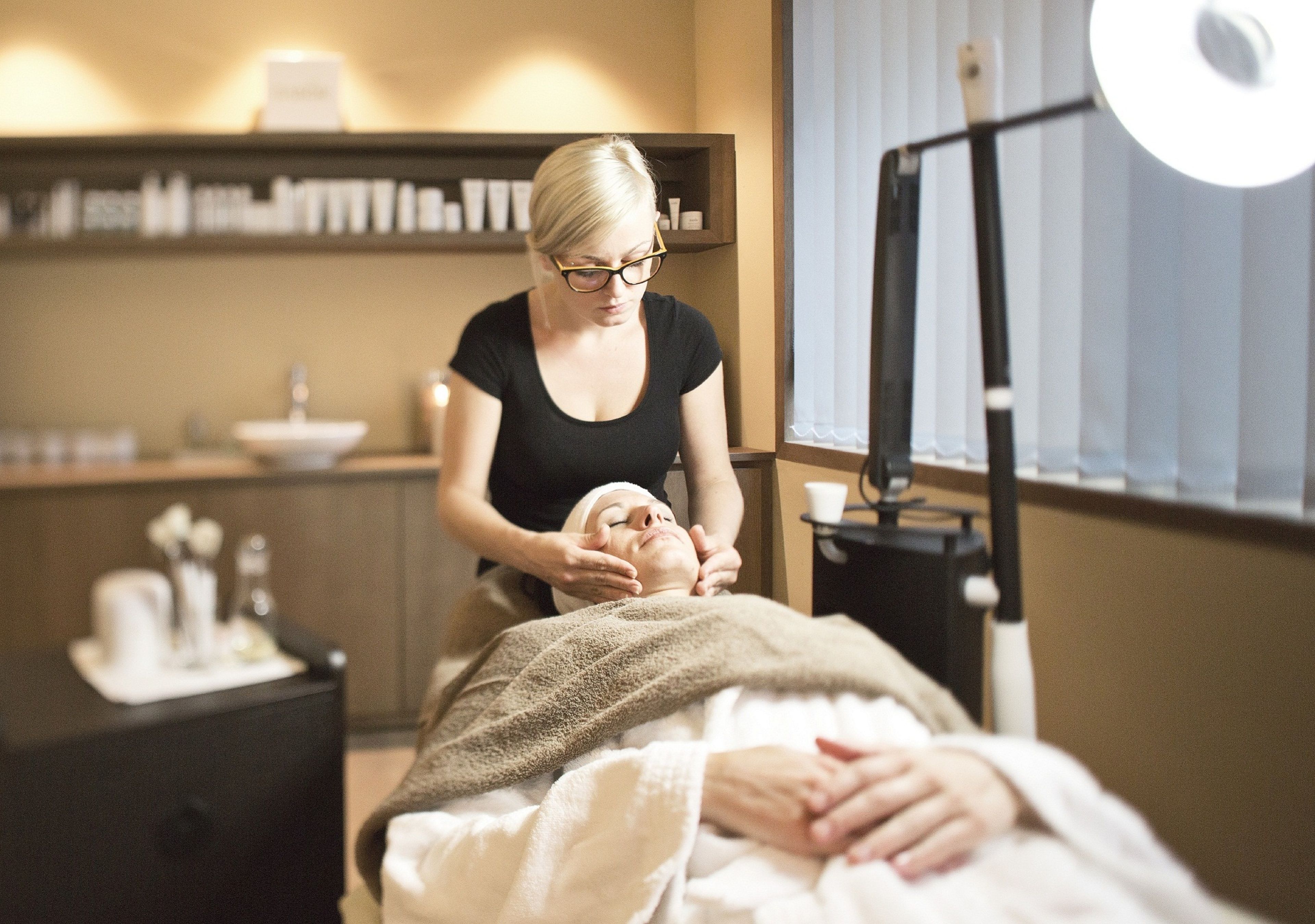 A person receiving a facial treatment in a room with a bed, a nightstand, and a shelf with toiletries.